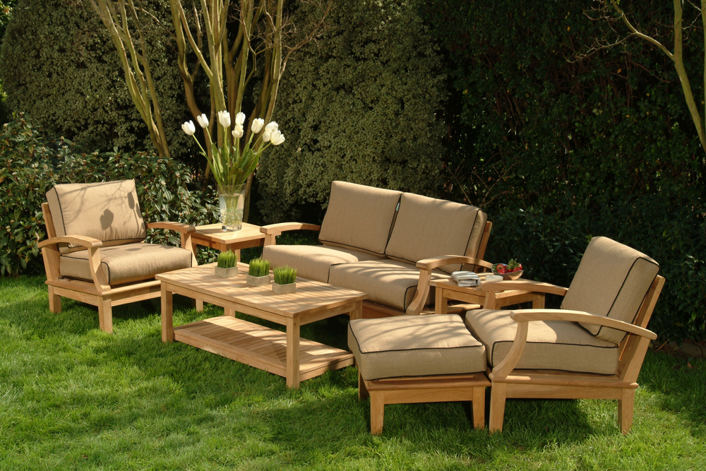 Conjunto de muebles de jardín de madera con cojines beige, compuesto por un sofá, sillones individuales, una mesa de centro y mesas auxiliares, ubicado sobre césped con un fondo de árboles y arbustos, creando un ambiente acogedor y elegante.